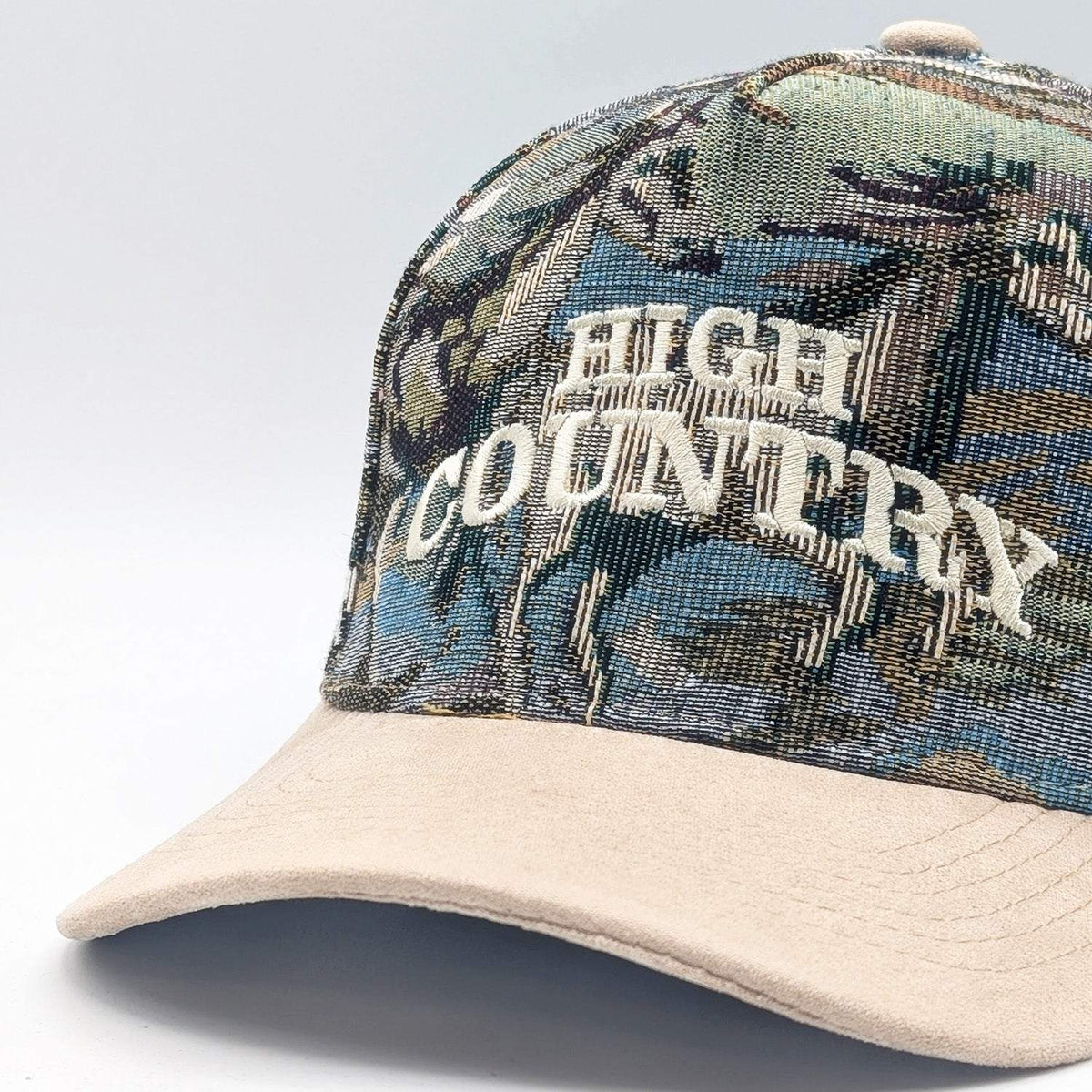 High Country Snapback with a blue, green, and brown jacquard countryside-patterned crown, cream embroidery, and tan suede brim.